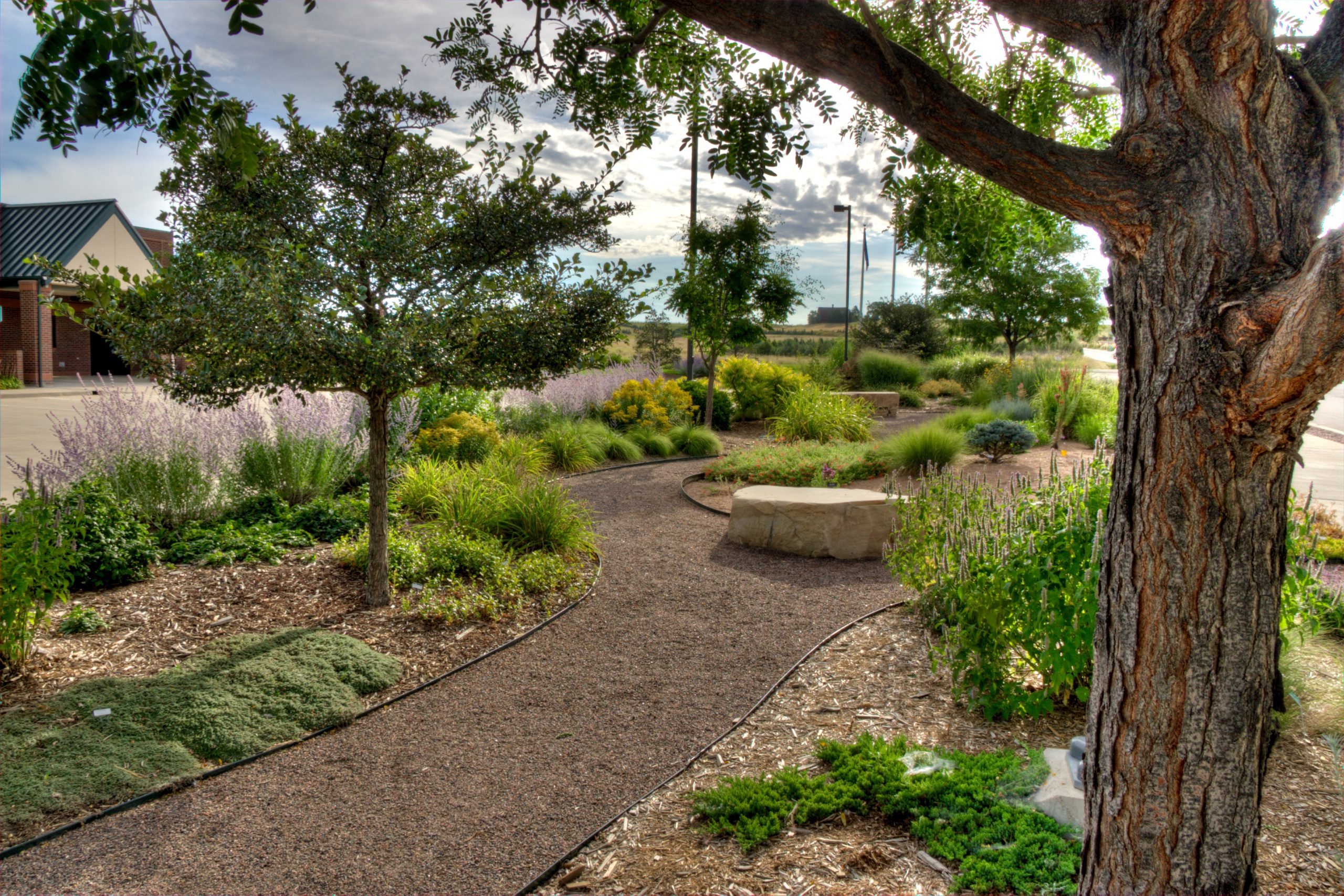 A gravel path shows the walk-through demonstration garden at Fire Station 5 with green trees and shrubs and the fire station building in the background.