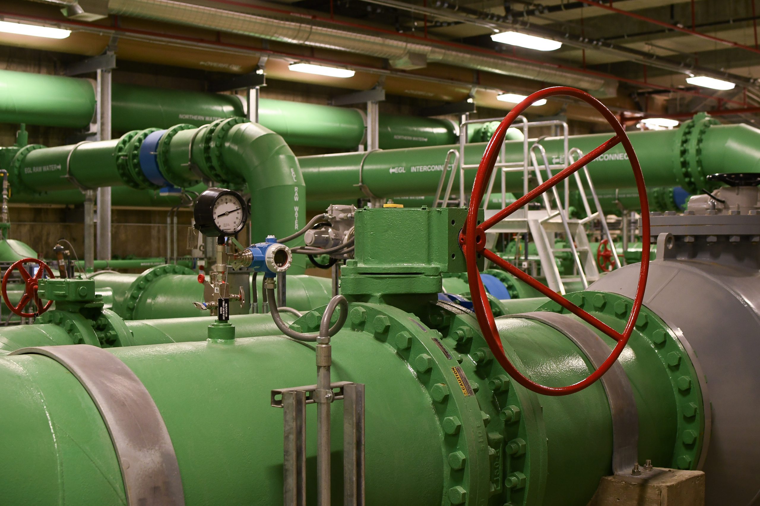 Large, green metal pipes in an indoor facility with large red wheel and measuring gauges.
