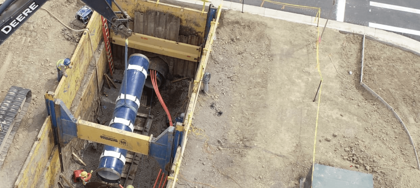 Aerial view of dirt construction site with an exposed blue underground pipe. There is an excavator on the left, and a street on the upper right.