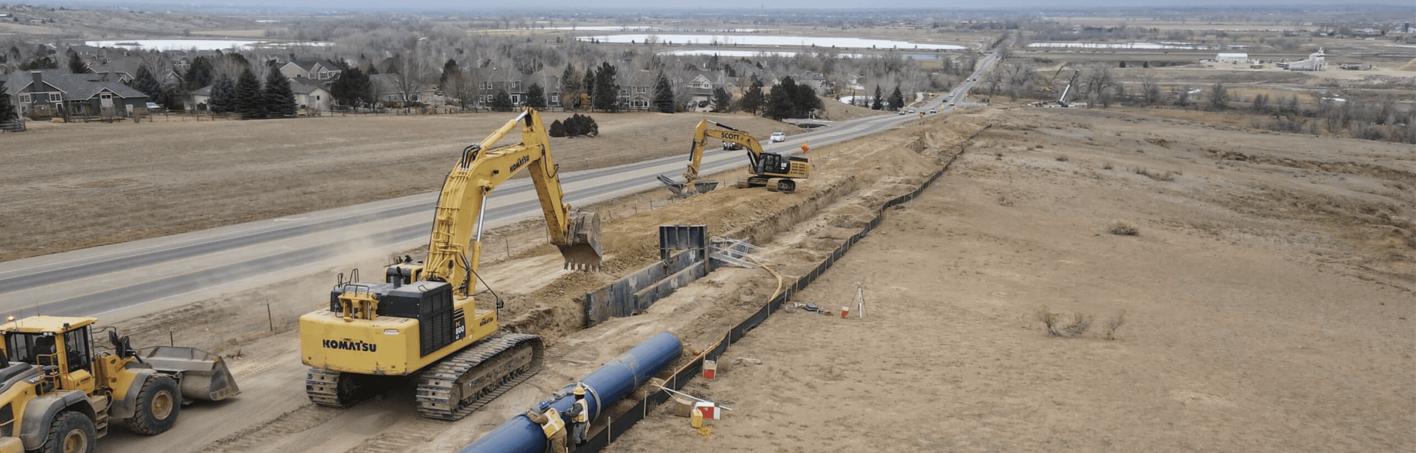 Dirt construction site with two yellow excavators digging into the ground. On the left there is a two-lane road.