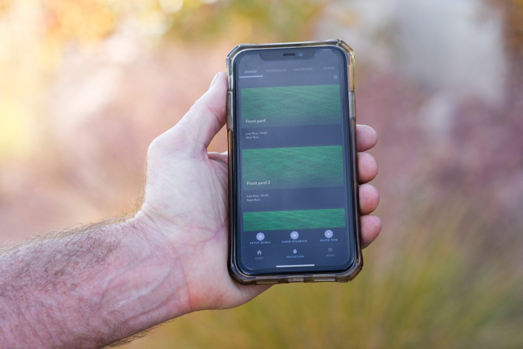 An outstretched hand holds a smartphone displaying the rachio water app.