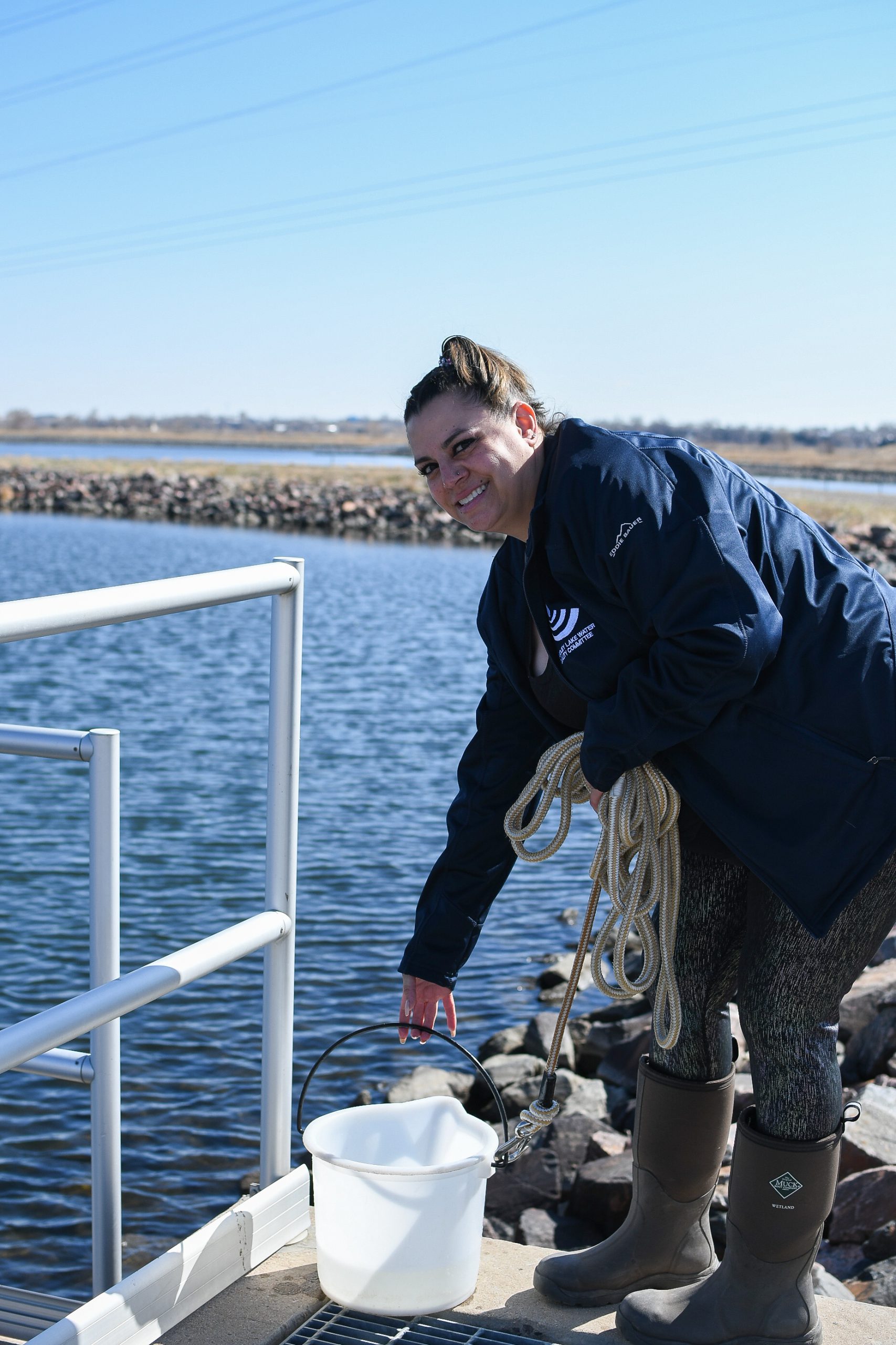 > Person with dark blue jacket, grey leggings and waterproof boots holding a rope and bucket a platform over a Colorado reservoir.