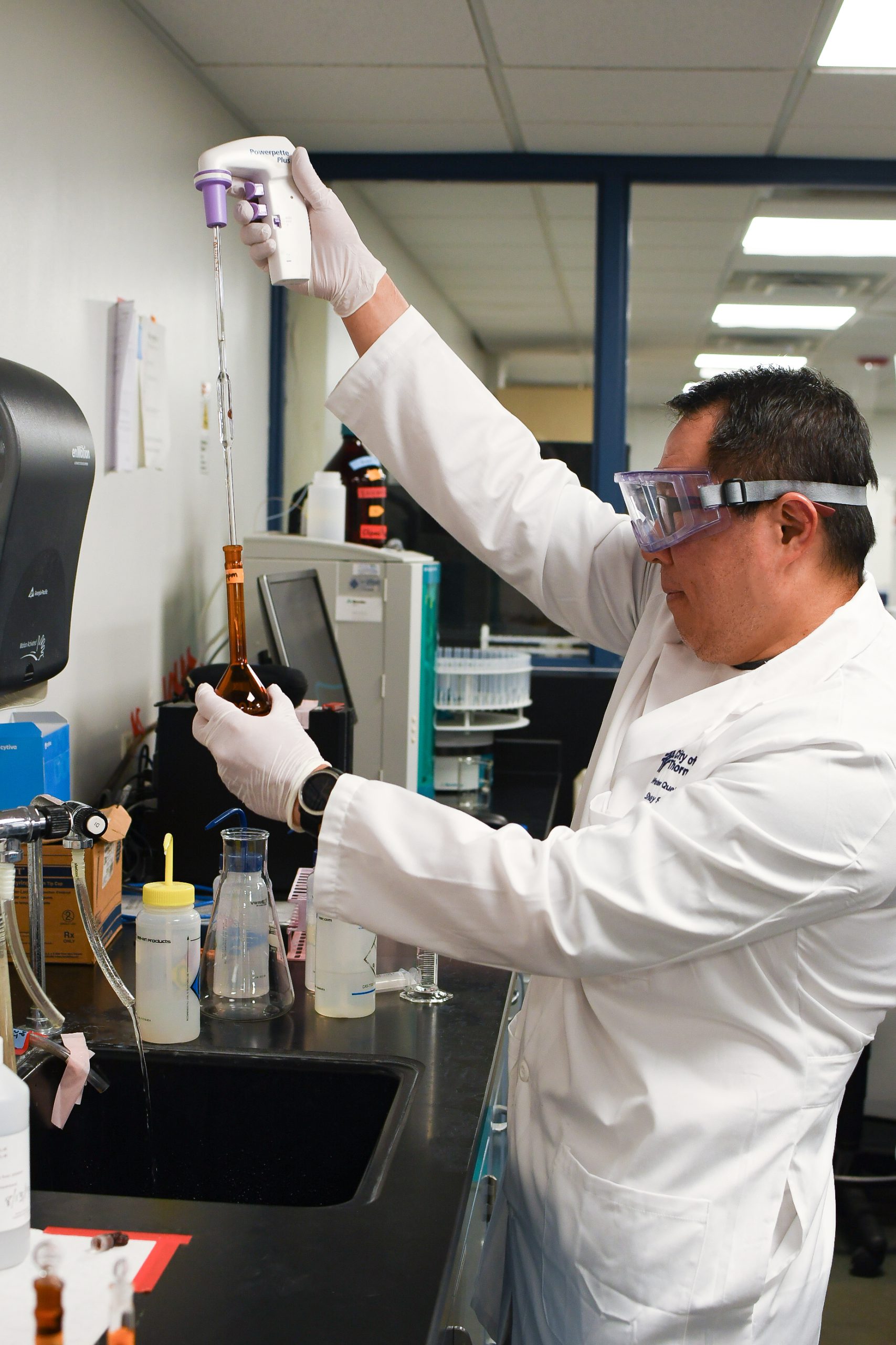 A man with dark hair wearing a lab coat and safety glasses uses a pipet to fill a sample from a brown tube.