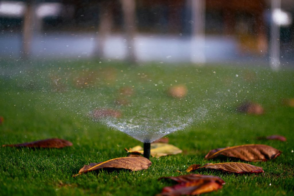 Small sprinkler head spraying water on green grass with fallen orange and brown leaves.