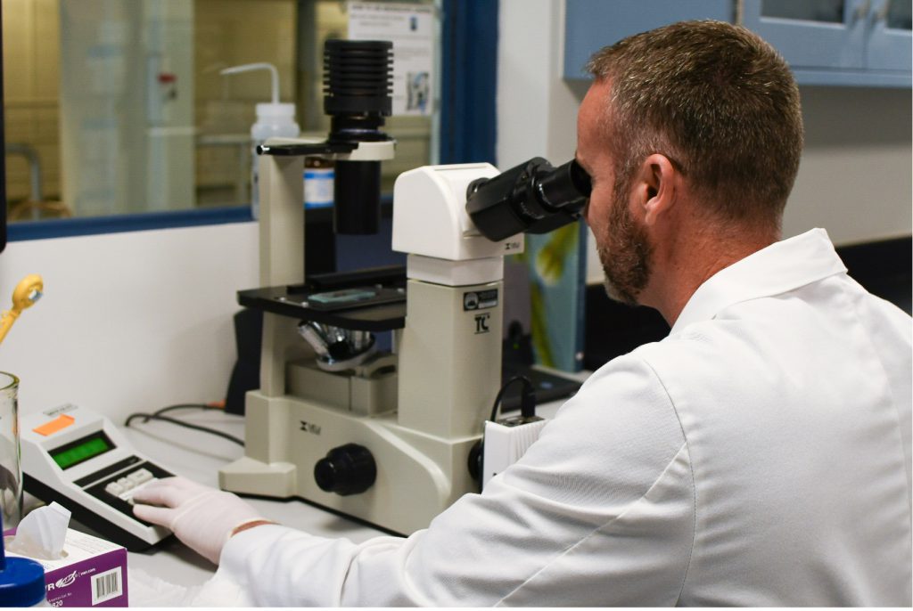 Man in a white lab coat and white rubber gloves looking through a microscope.