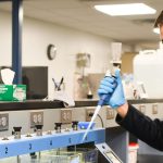 Person in grey jacket and safety goggles testing water samples at a water quality treatment plant.